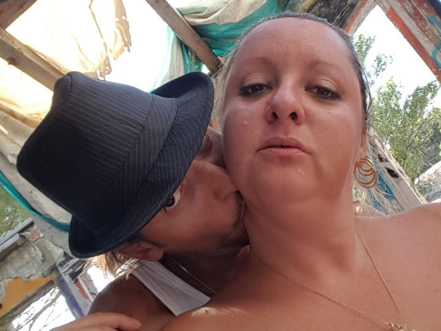 CplCokins with a chubby face, wet skin, and gold earrings, has a guy with a black hat kissing her neck. His face is partially hidden. She looks unimpressed.