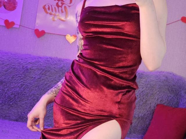 ShellyLove69 in a shiny red velvet dress, lifting it to reveal her thigh. Tattoo on her right arm, her hand pulling the dress up. Soft purple light.