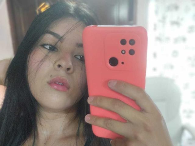 SalomeDias with black hair, pink lipstick, and a nose ring, holding a pink iPhone, taking a selfie, looking slightly bored. Her tits are not visible.