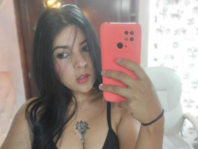 SalomeDias with black hair, pink phone, nose ring, and key tattoo on chest, holding phone to mirror, wearing black top, showing tits, and pink lips, in selfie.