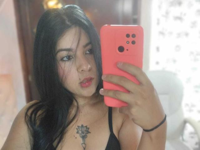 SalomeDias with long black hair, pink phone, nose ring, and black top, taking a selfie, showing her tits and a key tattoo on her chest.