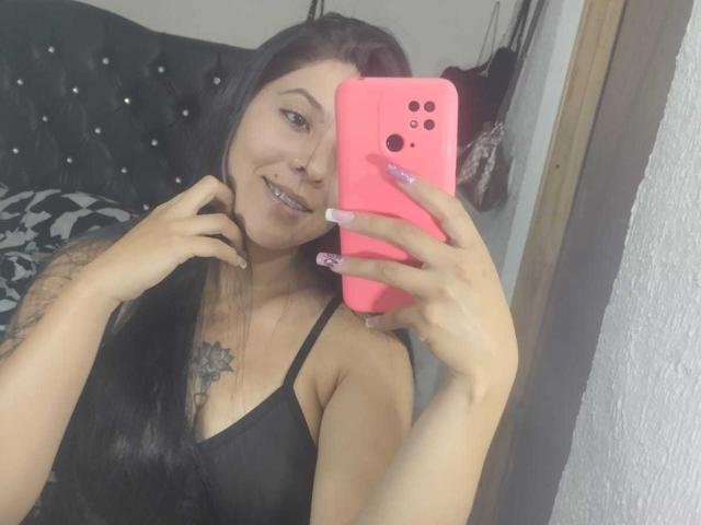 SalomeDias with long black hair, smiling, holding a pink phone, showing tattoos, wearing a black spaghetti strap top, reflecting in a mirror. Her tits are small and she has a nose ring.