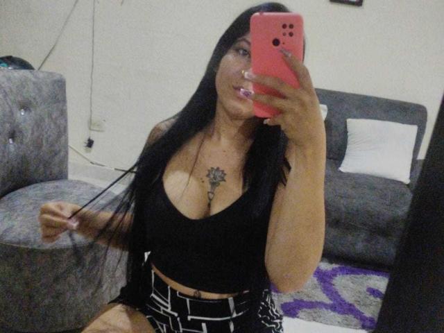 SalomeDias with long black hair, wearing a black tank top showing big tits and a tattoo above her cleavage, holding a pink phone, sitting in a grey room.