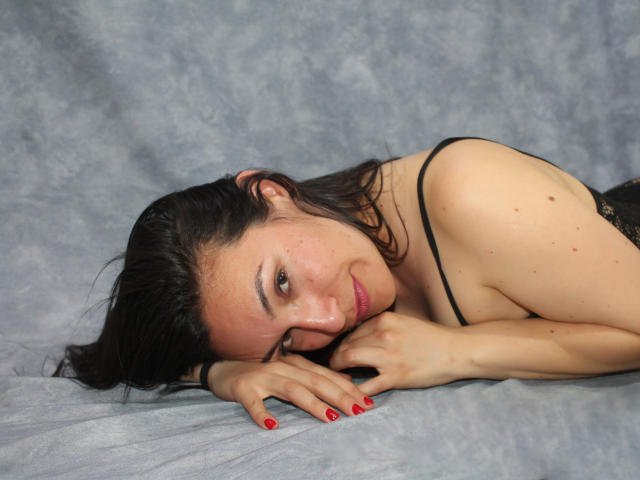 IvannaTish, com cabelo escuro, deitada de lado, vestindo lingerie preta, com esmalte vermelho, sorrindo para a câmera, mostrando seios pequenos, pele macia e um rosto bonito..