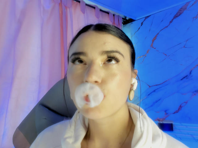 JasmineHypery, avec ses cheveux foncés, sa chemise blanche et son maquillage bleu pour les yeux, a une grosse bulle de substance gélatineuse rose-blanche dans la bouche, et porte des écouteurs blancs, dans une pièce très éclairée..