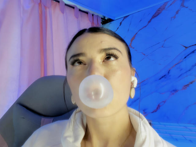 JasmineHypery, con cabello oscuro, soplando una burbuja blanca, vistiendo una camisa blanca, con un fondo azul y rosa, con auriculares blancos y mirando hacia arriba con largas pestañas..
