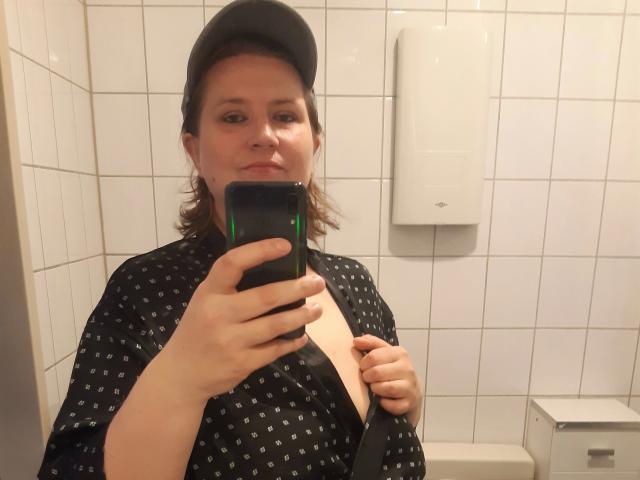 Uma mulher atraente, com cabelo castanho, usando um boné preto e um robe estampado, exibindo seus seios nus em um banheiro com azulejos brancos, segurando um celular e uma alça..