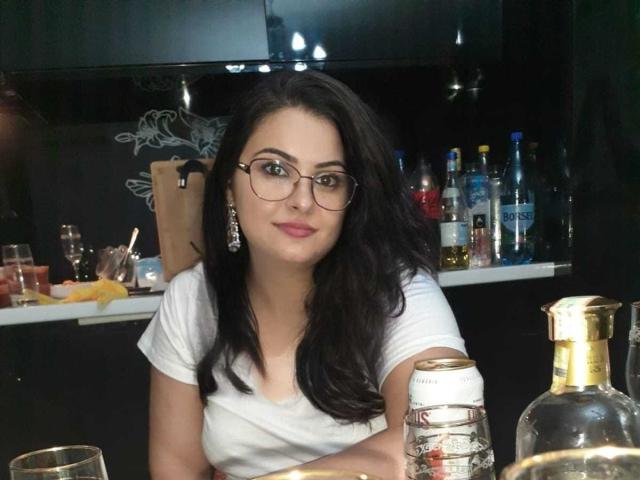 KathyKrystal, con cabello negro largo, gafas, camiseta blanca, sentada en la barra, mirando a la cámara, con senos pequeños, aretes, botellas y latas en la barra frente a ella, con una ligera sonrisa..