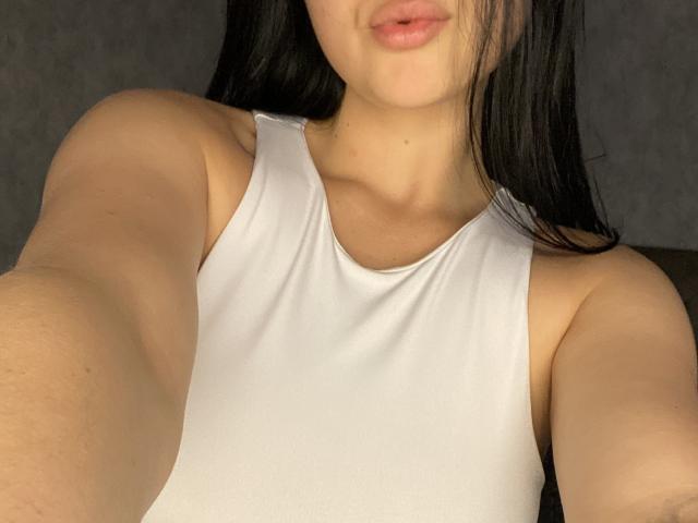 Le visage et le haut du corps d'Olieri sont visibles, elle porte un haut blanc sans manches, avec des seins apparents, des lèvres d'un rose clair et des cheveux noirs et lisses, et elle prend un selfie avec son bras droit..