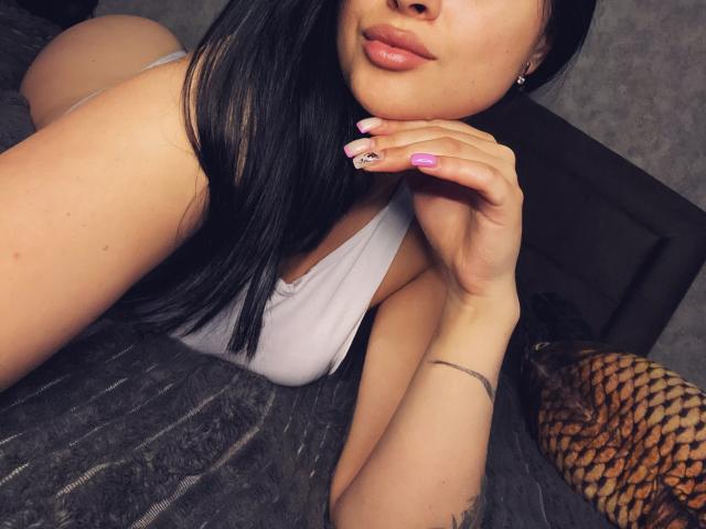 Olieri, lying on a bed, wearing a white tank top, showing her ass, with black hair, pink nails, and a silver bracelet, looking down at her hand.