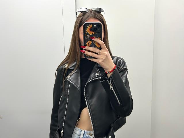 Karish, com cabelos castanhos longos, jaqueta de couro preta, unhas vermelhas, capa de celular com girassóis e uma blusa preta que expõe seus seios, está em pé em um quarto com paredes brancas..
