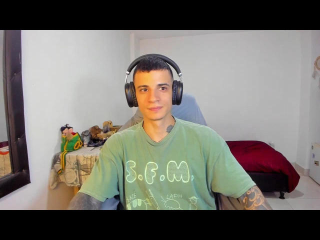 BaloForYou, con cabello castaño corto, usando auriculares negros, una camiseta verde, tatuajes en los brazos, sentada en una habitación, sonriendo, tiene un pequeño tatuaje verde en el cuello, y lleva mangas grises..