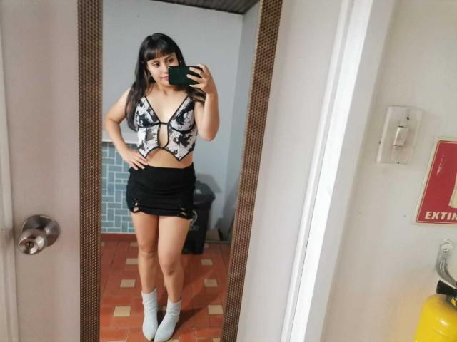 PaulaCeleste with black skirt, white socks, and floral bustier, showing her tits in a mirror selfie, holding a phone, standing in a tiled room, with a door.