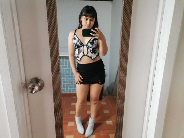 PaulaCeleste with black hair, bangs, white floral crop top with black straps, black mini skirt, white socks, standing in front of mirror, holding phone, showing her tits.