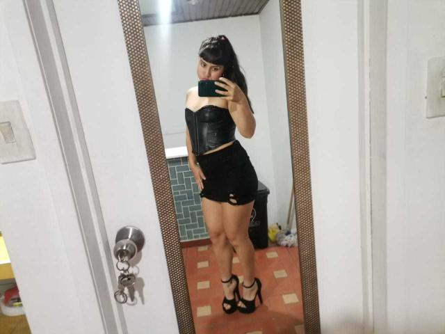 PaulaCeleste in a black leather corset and black mini skirt, standing in high heels, taking a mirror selfie with a black phone, showing her ass and toned legs.