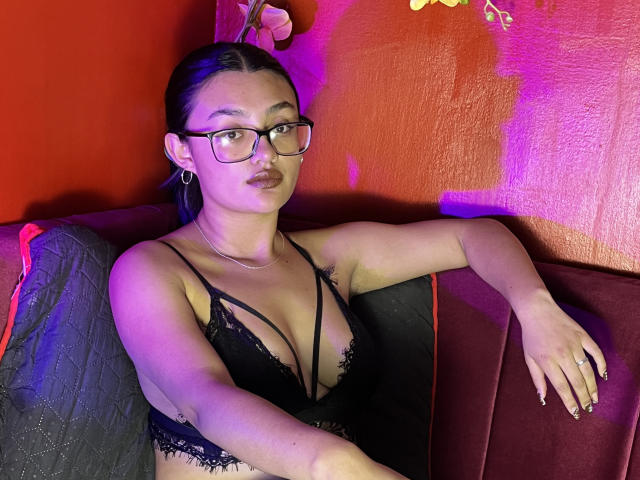 EmiilySwett with glasses, black lace lingerie, showing her tits, sitting on a red and black couch, arm resting on the back, purple and pink lighting.