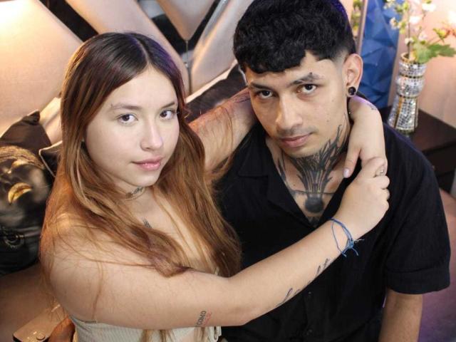 BelialAndLilim with long brown hair, small tits, arm tattoo, and a black shirt, hugs BelialAndLilim with curly black hair, tattoos, and a black shirt. Both look at camera.