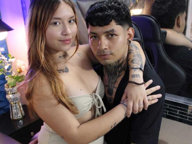 BelialAndLilim with long brown hair, small tits, and tattoos, hugs a tattooed man with short black hair, black shirt, and ear gauges. Both look at the camera.