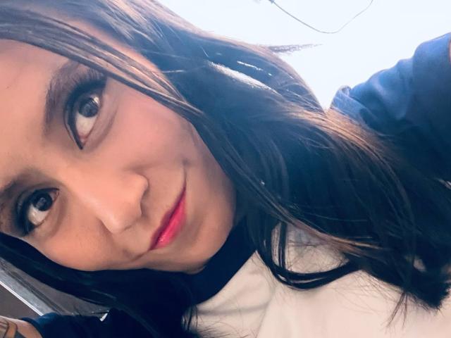 EviieGothel has long brown hair, brown eyes, and a slight smile with pink lipstick. She's wearing a black and white shirt, looking up with a cute, relaxed expression.
