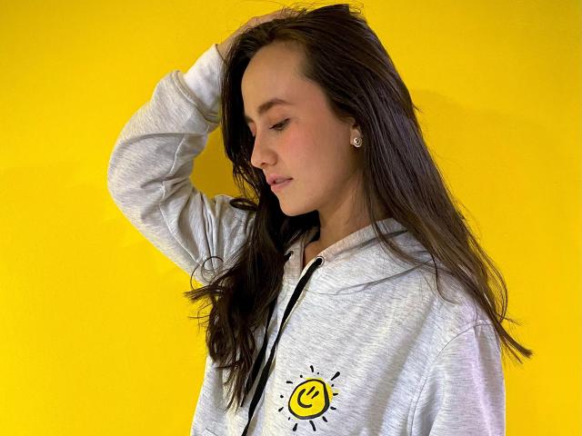 Lila69Hot with long brown hair, wearing a gray hoodie with a yellow smiley logo, hand in hair, looking down, yellow background. Her ass is not visible.