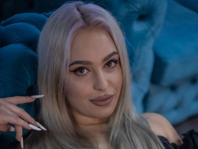 BiaKristal mit platinblondem Haar, braunen Augen und langen Wimpern, lächelnd mit matter rosa Lippenfarbe, trägt Schwarz und zeigt weiße Nägel, sitzt auf einem blauen, gepolsterten Sofa..