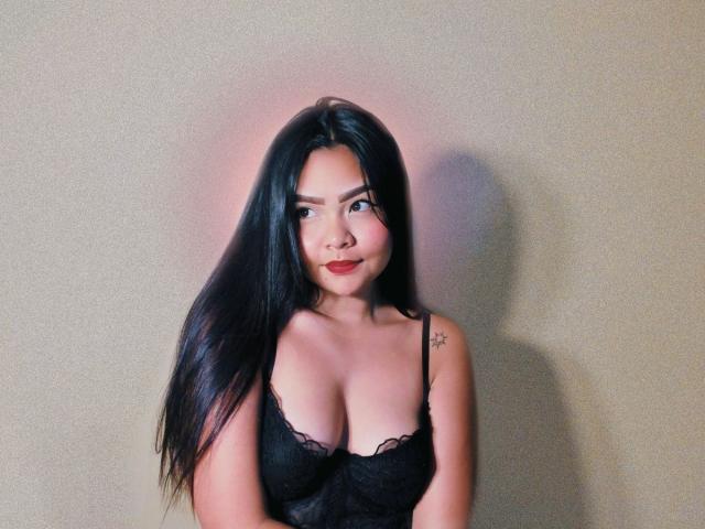 LovelyAnaX with long black hair, wearing a black lace bra, showing her big tits, a small star tattoo on her right shoulder, and a cute smile.