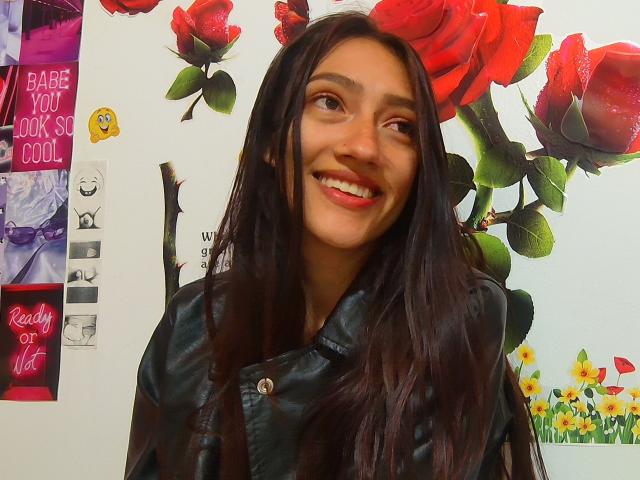 KiriamGomez, mit einem roten Lippenstift lächelnd, hat langes, dunkelbraunes Haar, trägt eine schwarze Lederjacke und blickt mit einem verspielten Ausdruck zur Seite, auf einem farbenfrohen Foto mit floralen Motiven..