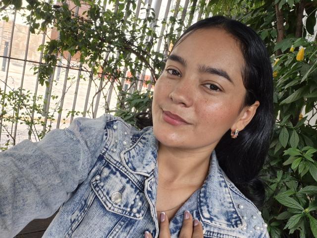 LilianaPink with medium brown skin, black hair, denim jacket, and subtle makeup, smiling slightly, wearing small earrings, holding her jacket collar, green leafy background, casual selfie.