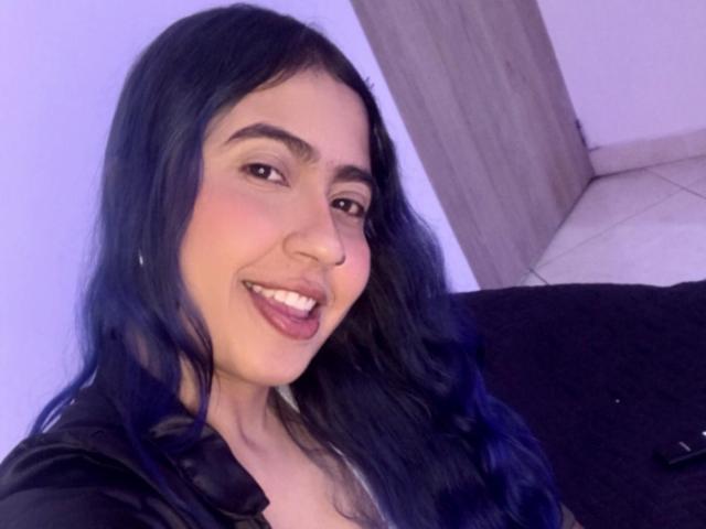 AriaJhonsson with long blue hair, smiling with her mouth open, showing teeth, wearing a black top, looking at the camera, with light skin and brown eyes.