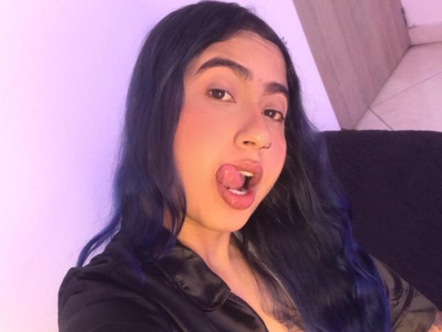 AriaJhonsson with blue hair, nose ring, and open mouth, tongue out, wearing a black shirt, looking at the camera, soft purple lighting, long wavy hair, slightly surprised expression.
