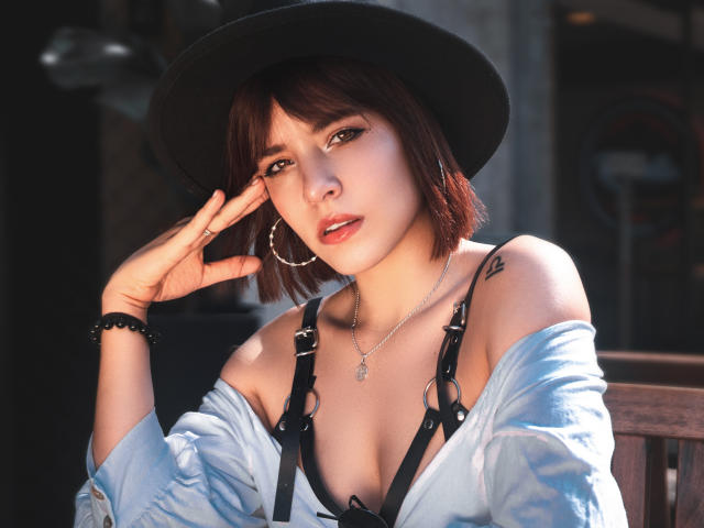 MilaFiore with short brown hair, black hat, and white shirt off-shoulder, showing small tits in black harness, hand near face, hoop earrings, black bracelet, tattoo on right shoulder.