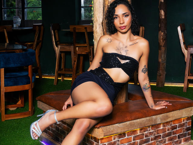 SilvanaXOwen with curly black hair, tan skin, tattoos, wearing a black strapless dress, sitting on a wooden table, showing her ass, with silver high heels.