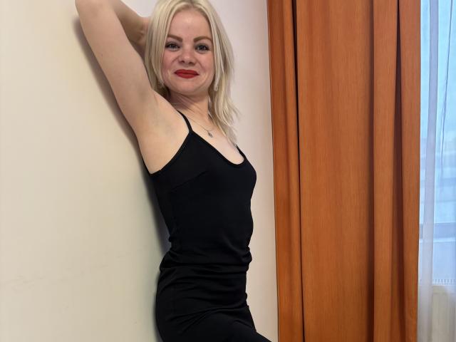 IrynaLyna mit blonden Haaren, rotem Lippenstift, schwarzem Kleid, Arm erhoben, an einer Wand lehnt, lächelt, zeigt kleine Brüste, schlank, helle Haut, orangefarbene Vorhänge, Fenster, Tageslicht..