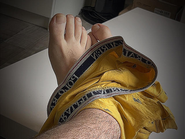 HombreDiscreto's hairy legs with yellow boxer shorts pulled down, exposing his bare feet, resting on a white table. Boxer shorts' waistband is visible, with "VANS" written on it.
