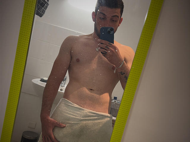HombreDiscreto with a towel covering his ass, shirtless, holding a phone in a mirror selfie, showing a tattoo on his right arm, in a bathroom.