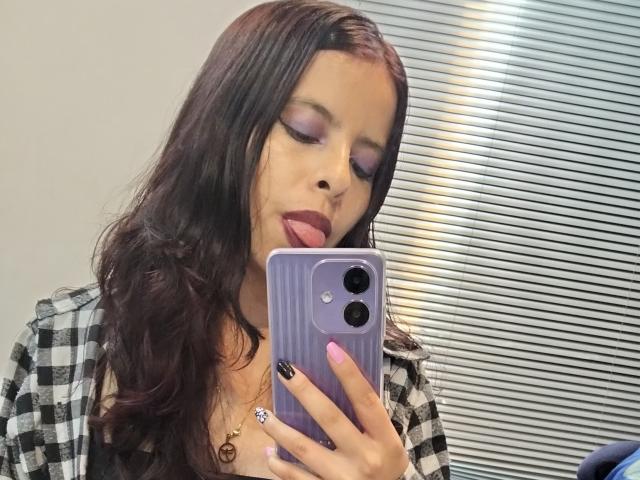 Julia Waine, con cabello largo y oscuro, sombra de ojos morada, lápiz labial rosa, lengua afuera, revisando el teléfono con una funda morada, uña negra, uña rosa, camisa de cuadros, collar, sosteniendo el teléfono en un selfie frente al espejo..