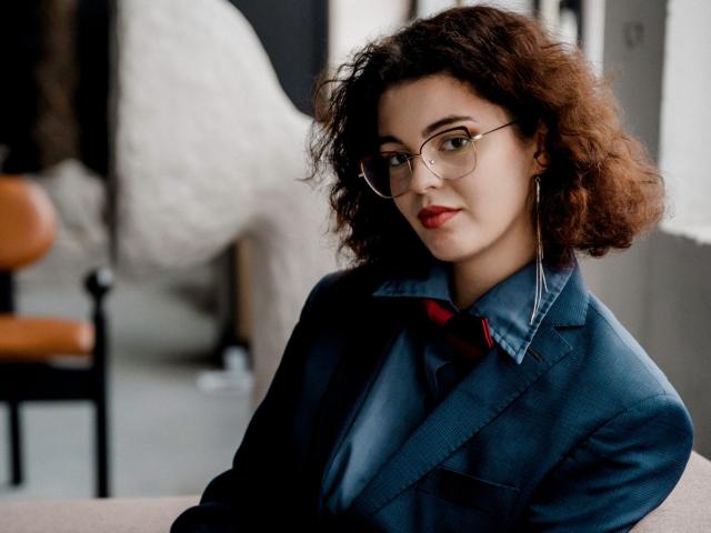 CurlyVixen, con cabello castaño rizado, usando gafas, una chaqueta vaquera azul, una camisa roja y aretes, tiene una apariencia sexy y segura, con lápiz labial rojo y una ligera sonrisa..