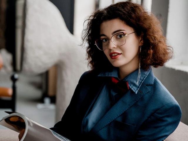 CurlyVixen with curly brown hair, wearing glasses and a blue shirt, holding a magazine, sitting, looking at camera with red lipstick, small hoop earrings.