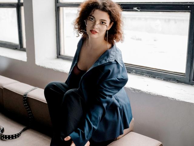 CurlyVixen with curly brown hair, glasses, and red lipstick, sitting on a bench wearing a blue shirt, black pants, and black underwear, showing her ass. 