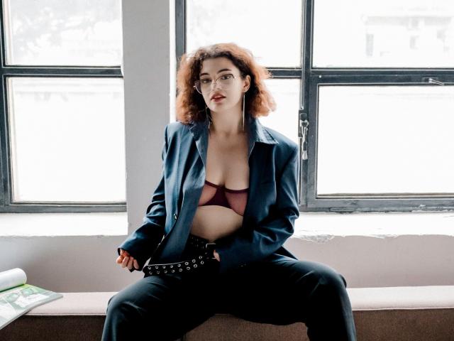 CurlyVixen with curly red hair, wearing a blue jacket, black pants, and a maroon bra, sitting with legs apart, showing her tits, in a bright room.