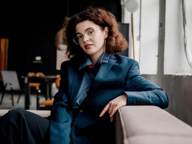 CurlyVixen with curly brown hair, wearing glasses, a blue suit jacket, and a red tie, sitting on a beige sofa, hand on her thigh, looking confident.