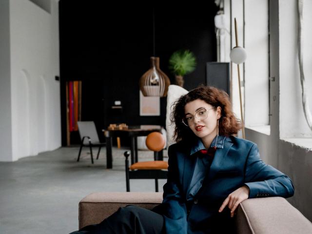CurlyVixen with curly brown hair, glasses, and red lipstick, wearing a blue suit and black pants, sits on a beige sofa, looking at the camera with a relaxed expression.