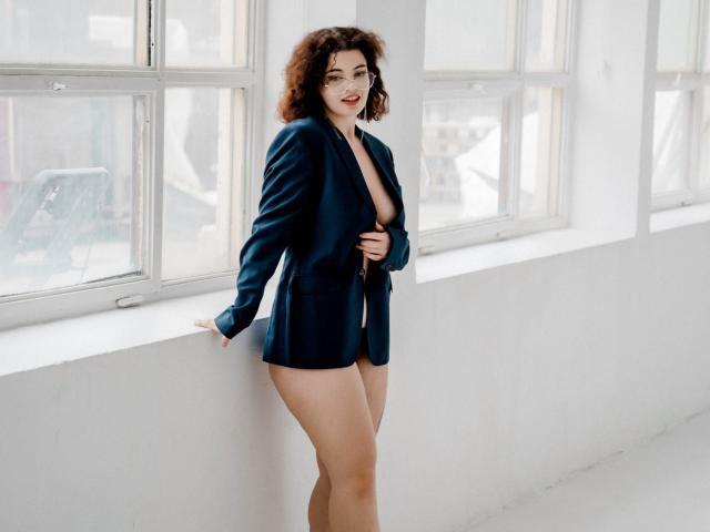 CurlyVixen with curly brown hair, wearing an open navy blue jacket, exposing her small tits and bare ass, standing by a window, smiling, in a bright white room.