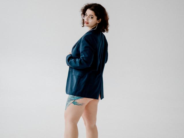 CurlyVixen stands in a dark blue shirt, exposing her bare ass and a teal tattoo on her right thigh, looking back with a slight smile. She has curly hair.