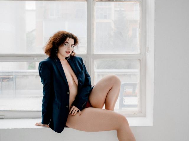 CurlyVixen with curly red hair, wearing an open navy blazer, exposing her large tits and bare ass, sitting on a windowsill, legs crossed, looking at the camera.