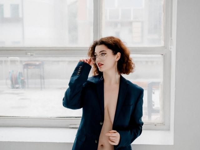 CurlyVixen with curly brown hair, wearing glasses and a navy blue blazer, open to reveal bare tits, standing in front of a window, touching her face.