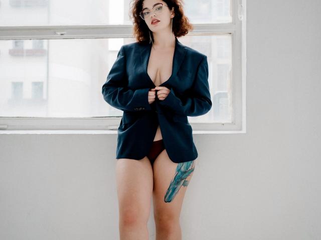 CurlyVixen with curly brown hair, wearing an unbuttoned dark blue blazer, exposing her big tits and red panties, has a green tattoo on her right thigh. Standing by a window.