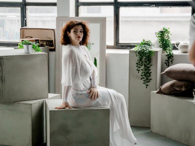CurlyVixen with curly red hair, white sheer dress, sitting on concrete block, hand on block, looking forward, soft light, green plant, brown pillow, large window in background.