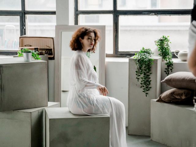 CurlyVixen with curly red hair, wearing a sheer white dress, sitting on a concrete block, looking over her shoulder, with green plants and brown pillows in the background.