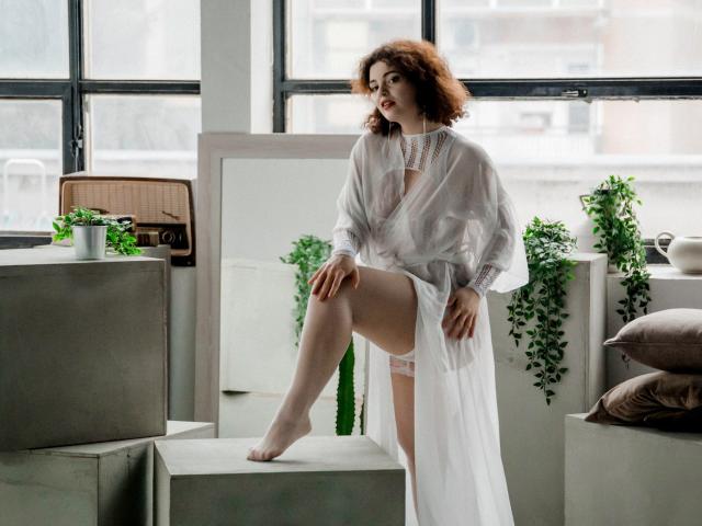 CurlyVixen with curly red hair, wearing a sheer white robe, exposing pink lace panties, foot on a gray cube, looking sideways, white lace necklace, indoor plant in background.